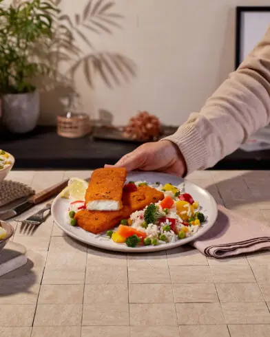 Človek si servíruje tanier s obaľovaným rybím filé značky FRoSTA, ryžou a zeleninou. Vidieť šalát, plátky citróna, vodu, karafu, rastliny a knihy.